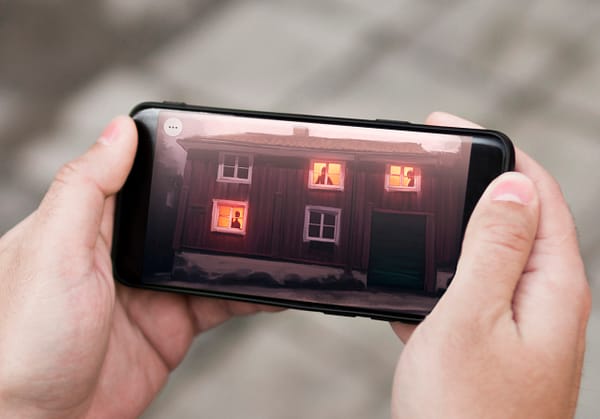 A close up photo of hand holding a smartphone. on the screen is an illustration of a haunted house with ghosts in the windows.