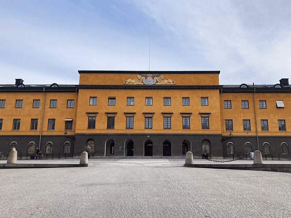 A photograph of Vitterhetsakademiens library in Stockholm. It is a three-story stone building painted a dark ockra color. In front is a large paved plaza.