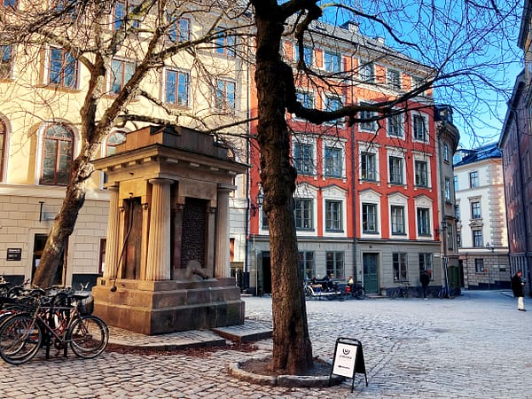 A photo of a little square in Stockholm's old town. In the foreground there is an old well, and in the background are a yellow and a red ston building against a clear blue sky