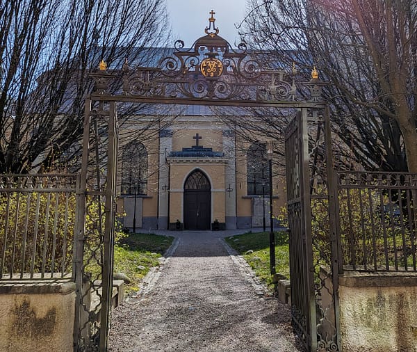 A photograph of the entrance to the German church cemetary. The yellow stone church building is surrounded by a black cast-iron fence with gold details.