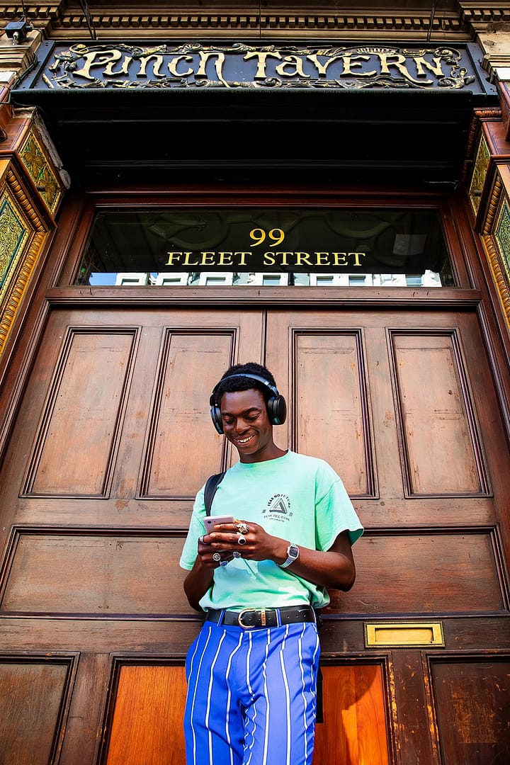 A young man is standing in front of a door. He is wearing headphones and looking at his phone, smiling.