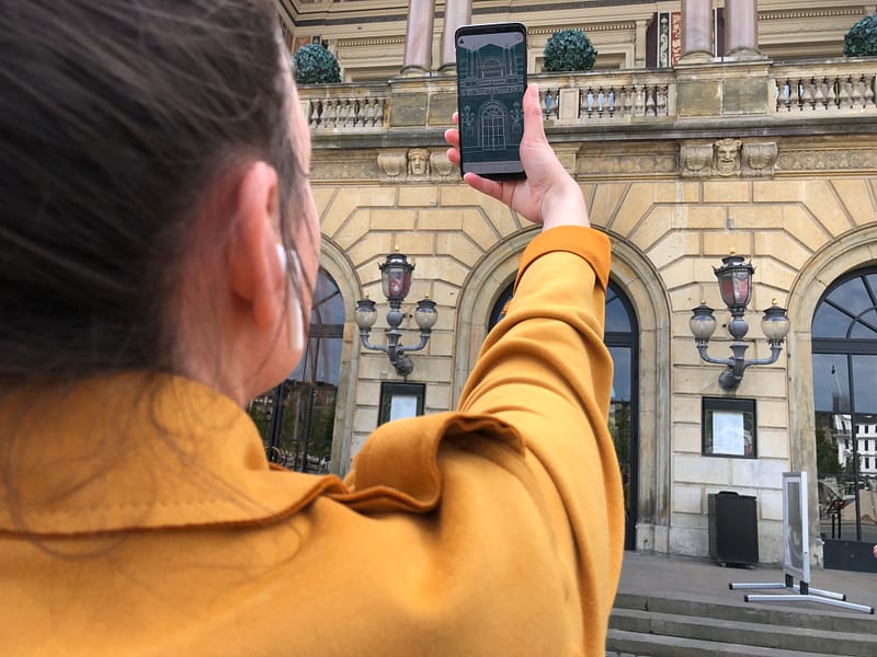 A woman in a yellow coat is holding up her smartphone, on the screen you can see a sketch of the building she is standing in front of